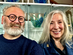 Head and shoulders of Alan (salt and pepper bear and short hair with black-rimmed round glasses) and Nicole (shoulder-length straight blonde/grey hair) Milligan smiling in front of a glass dish cabinet