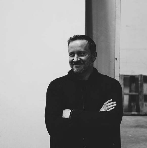 Waste-up black and white photo of James Culleton, wearing a long-sleeve, collared black shirt, short dark hair, crossing his arms, looking left with a closed smile